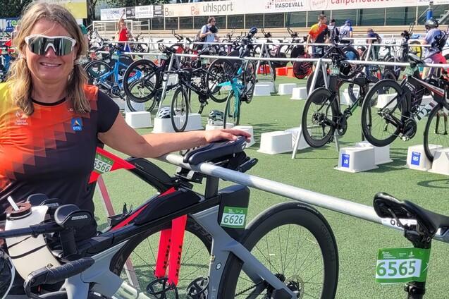 Frau mit Sonnenbrille steht neben ihrem Rennrad in einer Wechselzone beim Triathlon, mit weiteren Fahrrädern im Hintergrund.