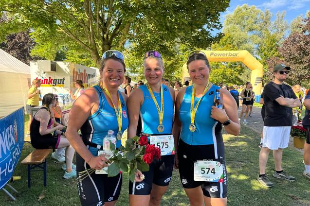 Drei Frauen in Sportkleidung halten Medaillen und Blumen, stehen lächelnd vor einer grünen Kulisse nach einem Wettkampf.