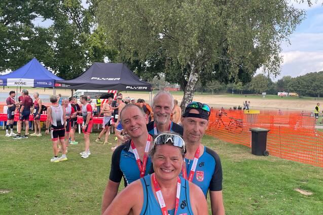 Gruppe von vier Triathleten mit Medaillen, nach einem Wettkampf im Freien, umgeben von Zuschauern und Pavillons.