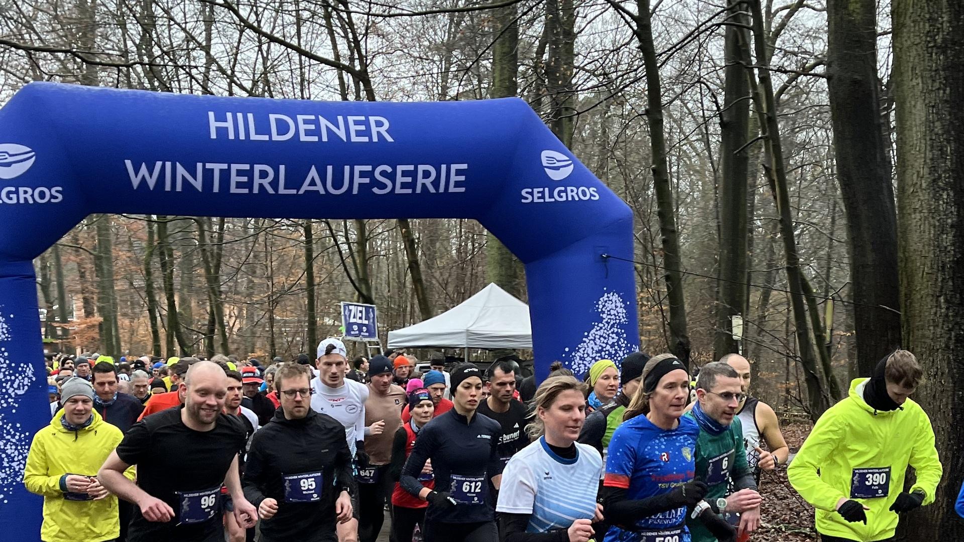Teilnehmer der Hildener Winterlaufserie starten unter einem blauen Tor in einem bewaldeten Gebiet.