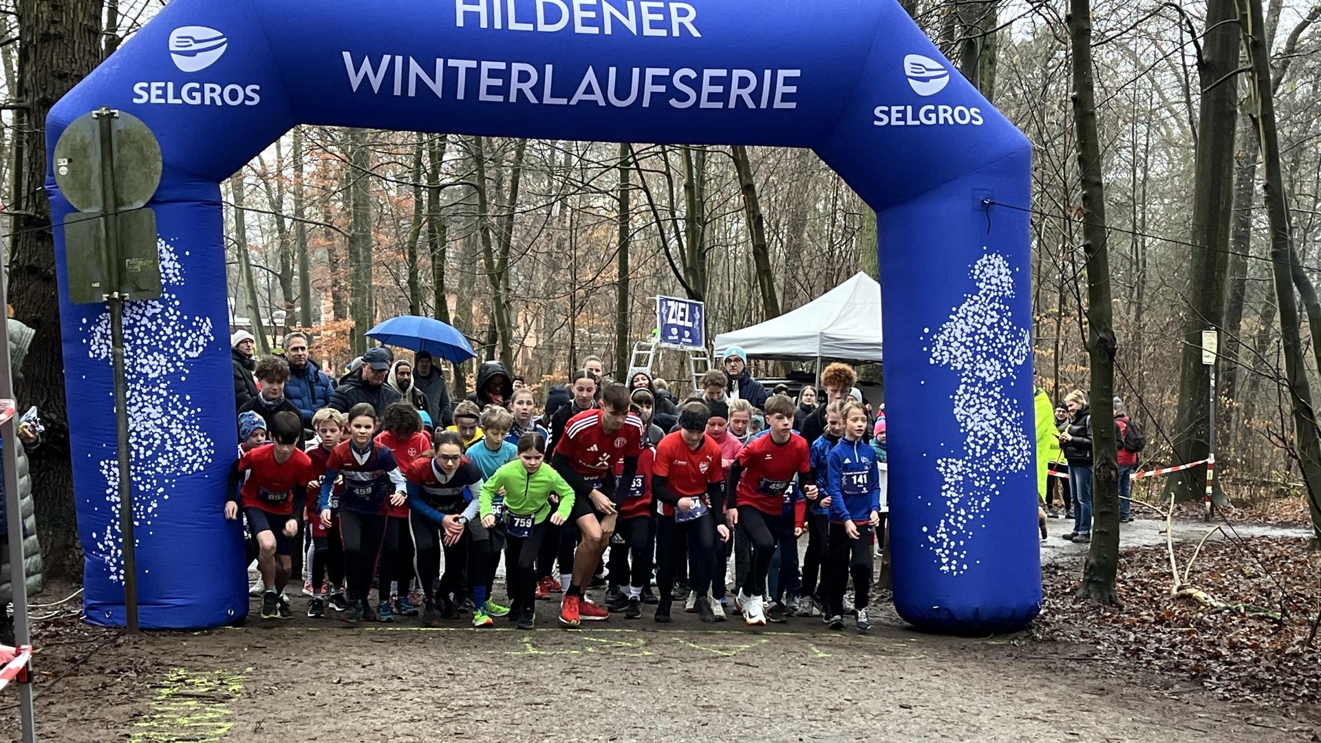 Startgruppe von Läufern unter einem blauen Bogen mit "Hildener Winterlaufserie" in einem Waldgebiet.