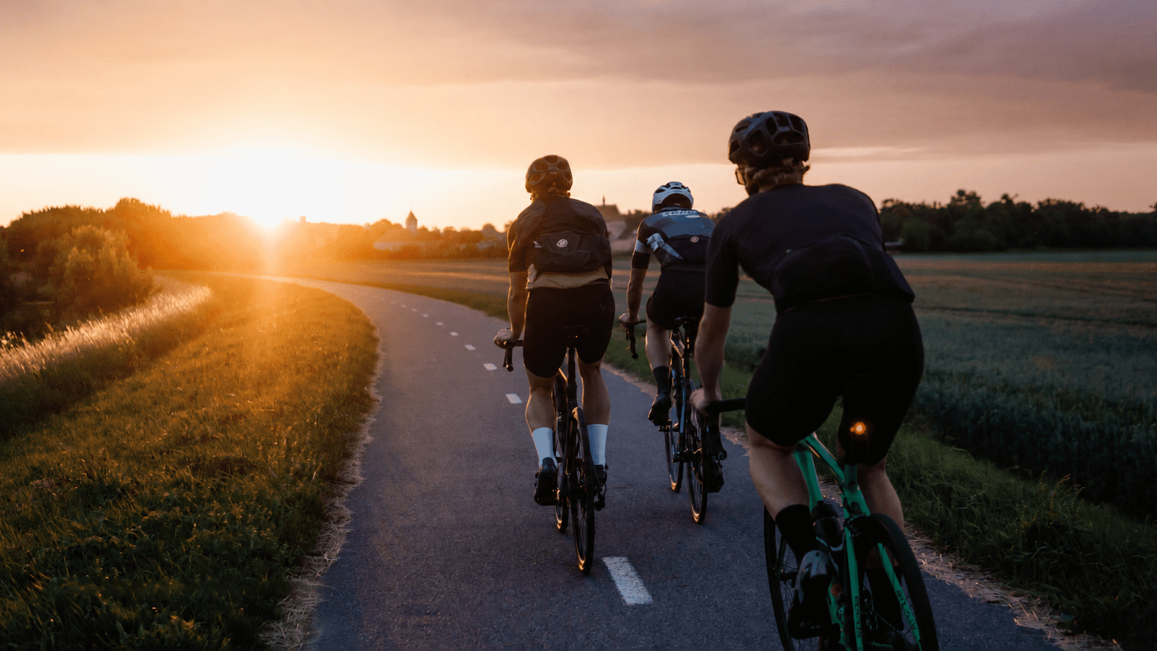 Drei Radfahrer fahren auf einem geschwungenen Radweg bei Sonnenuntergang durch die Landschaft.